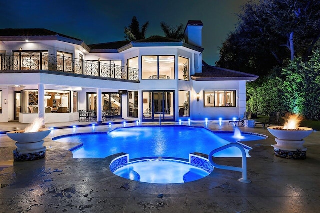 back of house at twilight featuring a patio, a balcony, a pool with connected hot tub, stucco siding, and a chimney