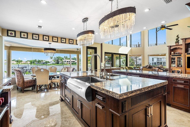 kitchen with pendant lighting, a sink, dark stone counters, a large island with sink, and ceiling fan with notable chandelier