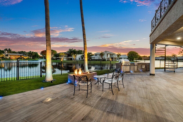 deck at dusk featuring an outdoor fire pit, a water view, fence, and a lawn