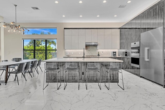 kitchen with built in appliances, light tile patterned floors, an island with sink, hanging light fixtures, and a breakfast bar