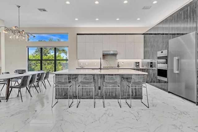 kitchen featuring light countertops, appliances with stainless steel finishes, modern cabinets, under cabinet range hood, and a kitchen bar