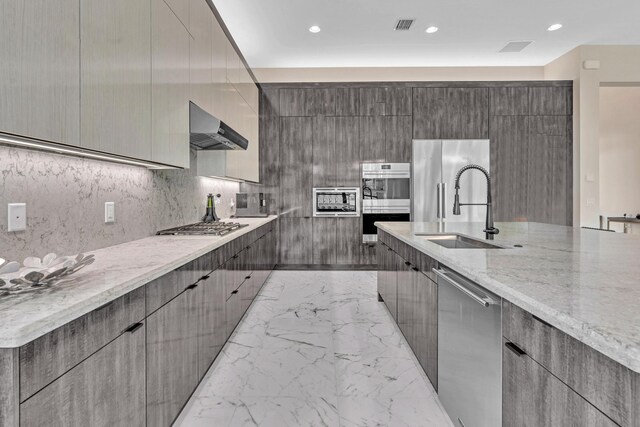 kitchen featuring light stone countertops, sink, wall chimney range hood, built in appliances, and decorative backsplash