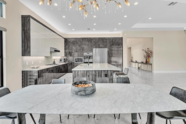 kitchen featuring backsplash, appliances with stainless steel finishes, a notable chandelier, a center island with sink, and a breakfast bar