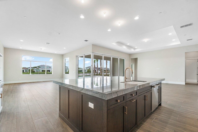 kitchen featuring dark brown cabinetry, sink, light stone counters, a tray ceiling, and a center island with sink