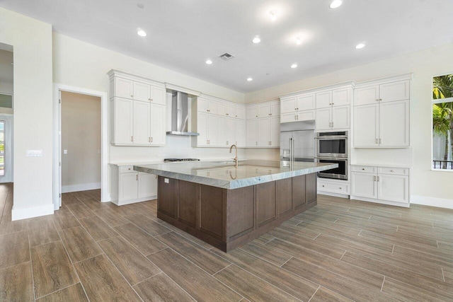 kitchen featuring a kitchen island with sink, wall chimney range hood, sink, white cabinetry, and stainless steel appliances