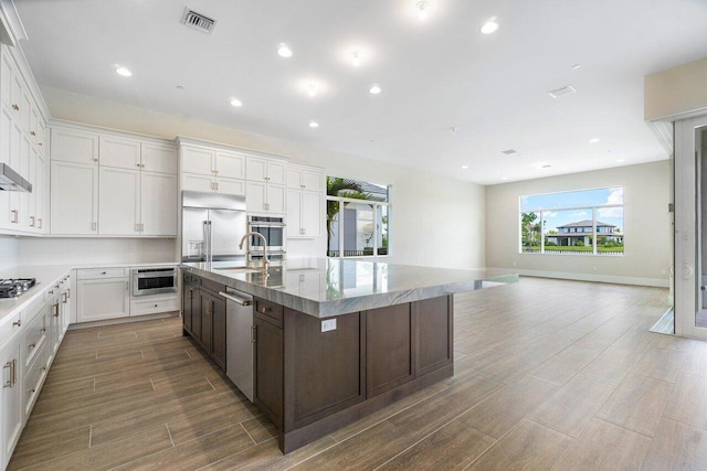 kitchen featuring light stone counters, a center island with sink, white cabinets, dark brown cabinets, and appliances with stainless steel finishes