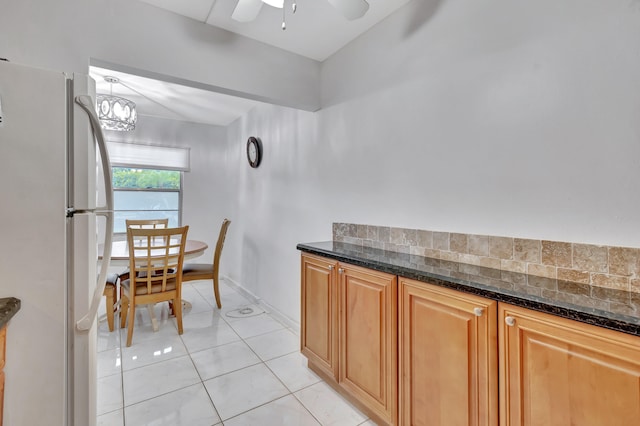 kitchen featuring white refrigerator, dark stone counters, ceiling fan with notable chandelier, and light tile patterned floors