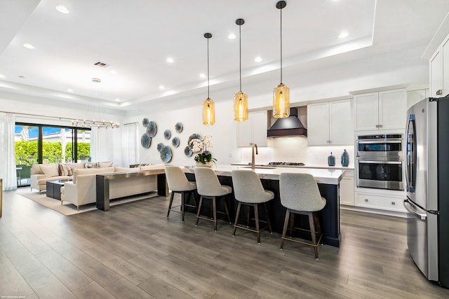 kitchen with premium range hood, dark hardwood / wood-style flooring, a tray ceiling, stainless steel appliances, and a center island with sink