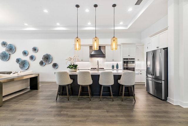 kitchen with premium range hood, hanging light fixtures, dark wood-type flooring, appliances with stainless steel finishes, and a kitchen island with sink