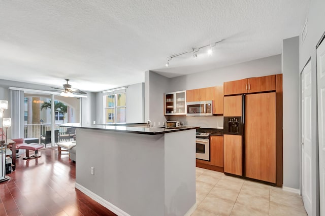 kitchen featuring brown cabinetry, a ceiling fan, dark countertops, appliances with stainless steel finishes, and a textured ceiling