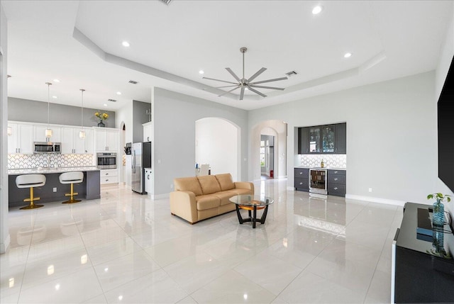 living room featuring baseboards, arched walkways, a raised ceiling, a ceiling fan, and wine cooler