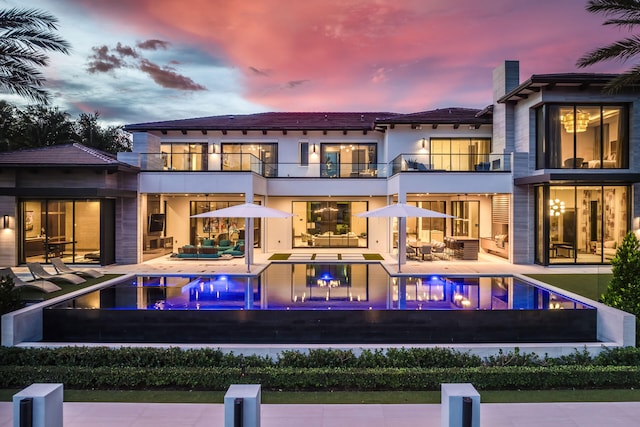 back house at dusk featuring outdoor lounge area, a swimming pool with hot tub, a balcony, and a patio area