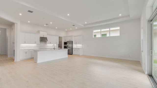 kitchen with light wood-type flooring, backsplash, stainless steel fridge, white cabinets, and a center island with sink