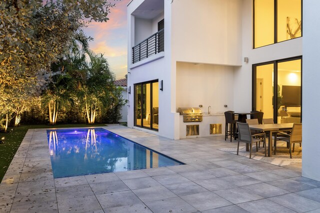 pool at dusk with exterior kitchen, grilling area, and a patio area