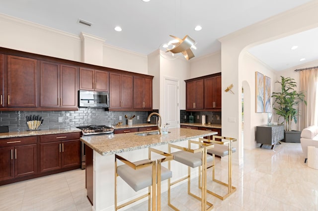 kitchen with light stone counters, ornamental molding, stainless steel appliances, a kitchen island with sink, and sink