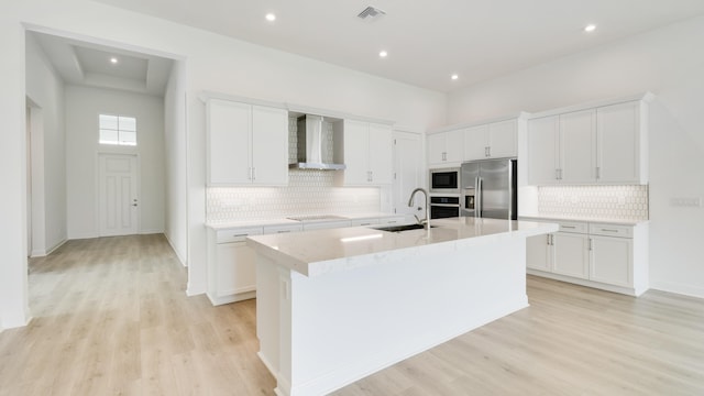 kitchen featuring appliances with stainless steel finishes, wall chimney range hood, a kitchen island with sink, and white cabinetry