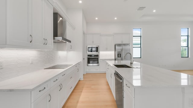 kitchen featuring a large island with sink, wall chimney range hood, sink, appliances with stainless steel finishes, and white cabinetry