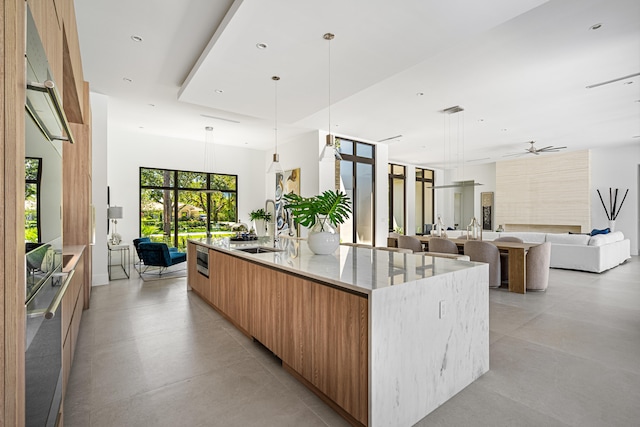 kitchen with decorative light fixtures, sink, light stone counters, an island with sink, and ceiling fan