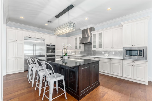 kitchen featuring built in appliances, white cabinets, a center island with sink, and wall chimney exhaust hood