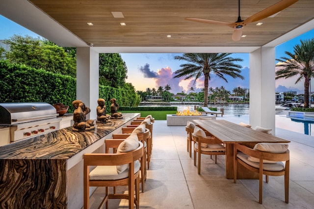 patio terrace at dusk with a water view, grilling area, area for grilling, ceiling fan, and a fire pit