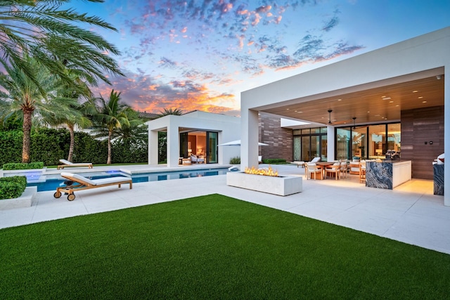 back house at dusk with a swimming pool with hot tub, a patio, a lawn, and a fire pit