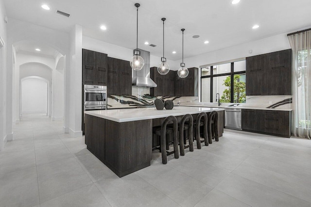 kitchen with tasteful backsplash, decorative light fixtures, a spacious island, appliances with stainless steel finishes, and wall chimney exhaust hood