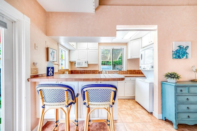 kitchen featuring a kitchen breakfast bar, stacked washing maching and dryer, light tile patterned floors, tile countertops, and white cabinets