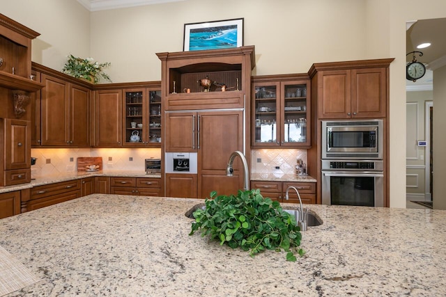 kitchen featuring built in appliances, light stone countertops, ornamental molding, brown cabinets, and glass insert cabinets