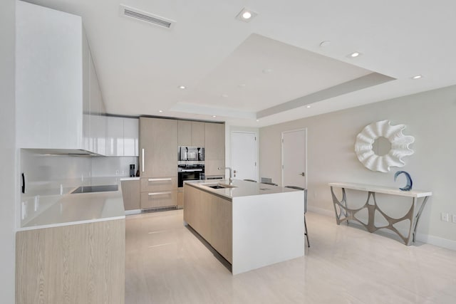 kitchen with sink, a center island with sink, black appliances, and a tray ceiling