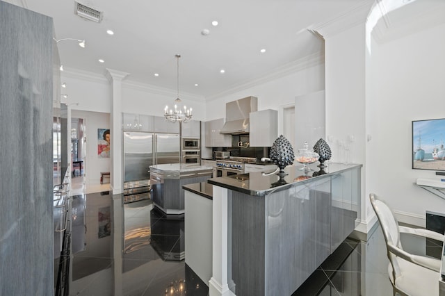 kitchen featuring kitchen peninsula, wall chimney exhaust hood, built in appliances, decorative light fixtures, and a breakfast bar area