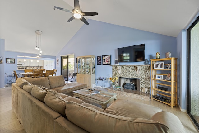 living room featuring high vaulted ceiling, light hardwood / wood-style floors, a stone fireplace, and ceiling fan