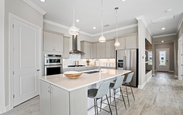 kitchen with sink, hanging light fixtures, a center island with sink, wall chimney range hood, and appliances with stainless steel finishes