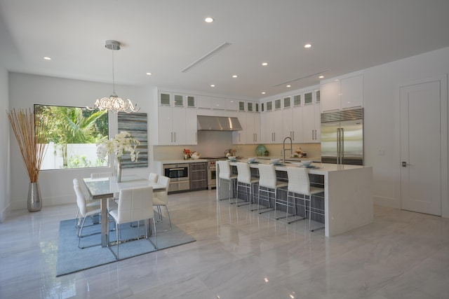 kitchen featuring decorative light fixtures, white cabinetry, stainless steel built in fridge, an island with sink, and wall chimney exhaust hood