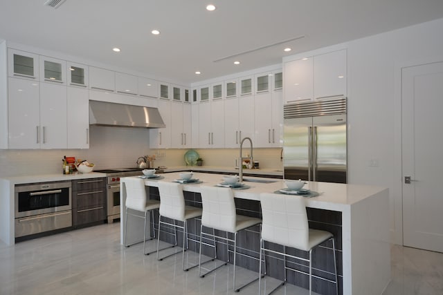 kitchen with high quality appliances, a kitchen island with sink, wall chimney exhaust hood, and white cabinetry