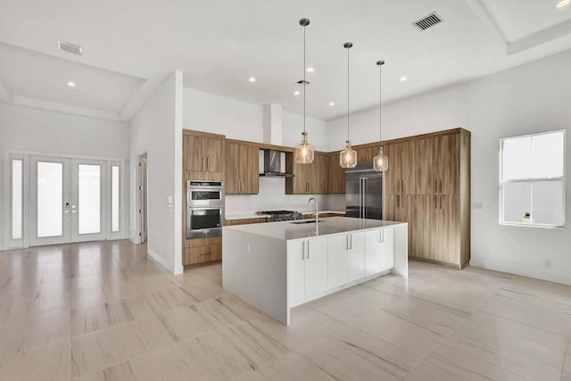 kitchen with wall chimney exhaust hood, sink, hanging light fixtures, stainless steel appliances, and a kitchen island with sink