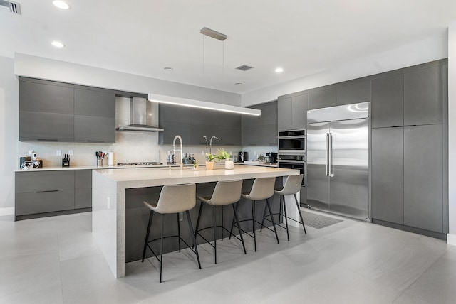 kitchen with backsplash, wall chimney range hood, a kitchen breakfast bar, an island with sink, and stainless steel appliances