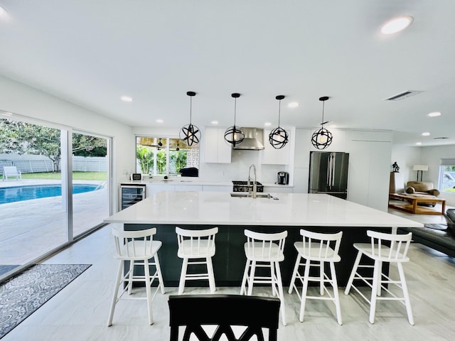 kitchen featuring white cabinets, decorative light fixtures, wall chimney exhaust hood, stainless steel fridge, and a large island