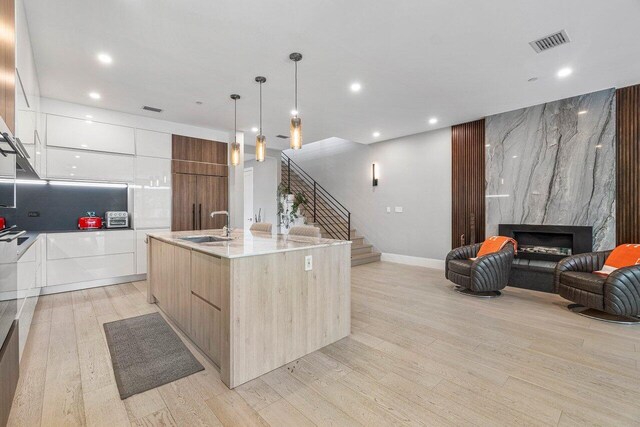 kitchen featuring light wood-type flooring, white cabinets, a kitchen island with sink, sink, and a high end fireplace