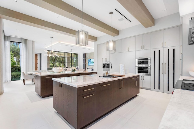 kitchen featuring a kitchen island with sink, dark brown cabinets, pendant lighting, light stone counters, and beamed ceiling