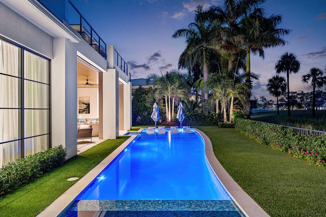 pool at dusk with an outdoor living space, ceiling fan, a patio, and a lawn