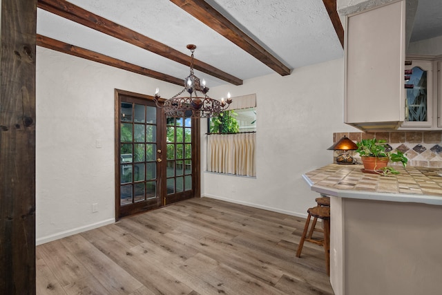 unfurnished dining area with a notable chandelier, a textured ceiling, french doors, beam ceiling, and light hardwood / wood-style floors