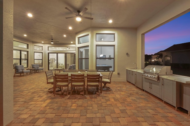 patio terrace at dusk featuring a grill, ceiling fan, sink, and an outdoor kitchen