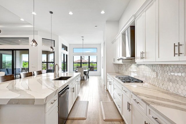 kitchen with decorative light fixtures, a kitchen island with sink, light stone counters, wall chimney range hood, and light wood-type flooring