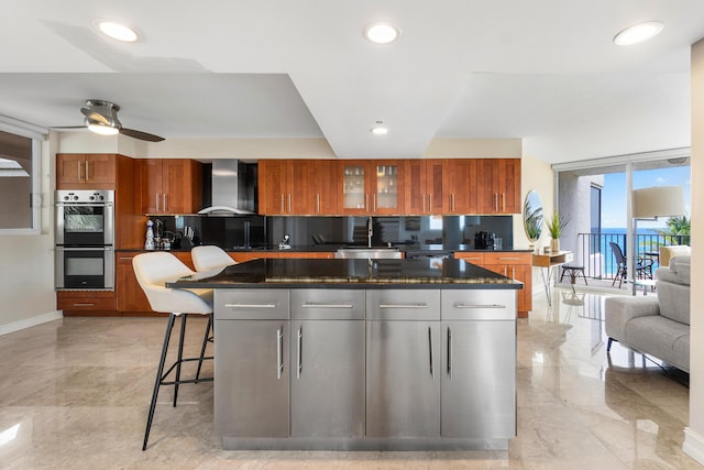 kitchen with a kitchen breakfast bar, ceiling fan, double oven, wall chimney range hood, and a center island