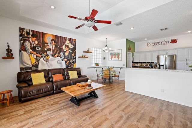 living room with a textured ceiling, light hardwood / wood-style flooring, ceiling fan with notable chandelier, and a raised ceiling