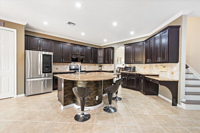 kitchen featuring light stone counters, range hood, stainless steel appliances, an island with sink, and dark brown cabinets