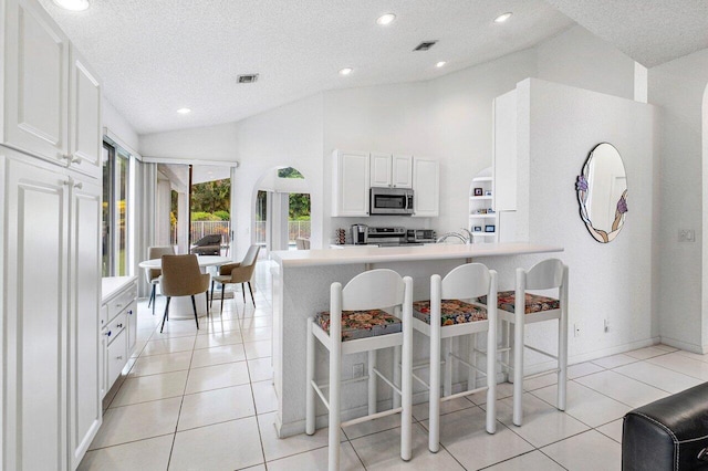 kitchen with a textured ceiling, light tile patterned floors, a kitchen bar, high vaulted ceiling, and white cabinets