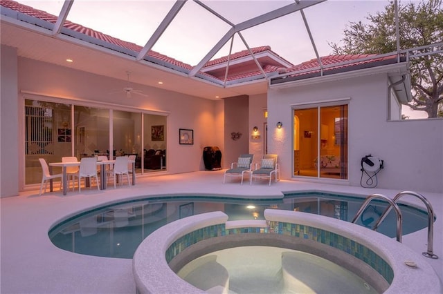 pool at dusk featuring an in ground hot tub, a patio area, glass enclosure, and ceiling fan