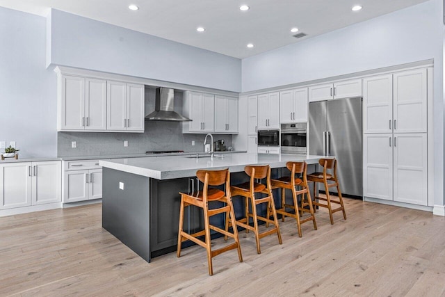 kitchen featuring wall chimney range hood, a kitchen breakfast bar, light hardwood / wood-style flooring, built in appliances, and a center island with sink