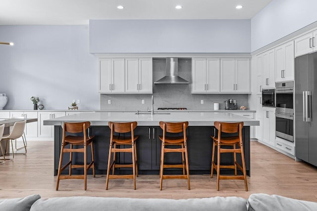 kitchen featuring wall chimney exhaust hood, an island with sink, appliances with stainless steel finishes, and light hardwood / wood-style flooring